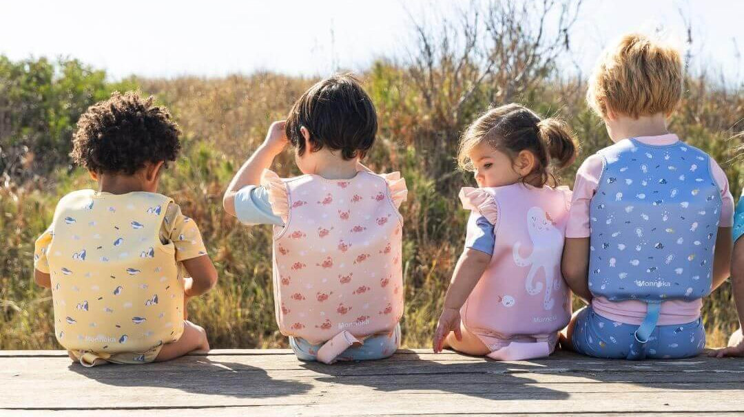Crianças usando coletes flutuadores coloridos Monneka, sentadas em um deck ao ar livre em frente à natureza.