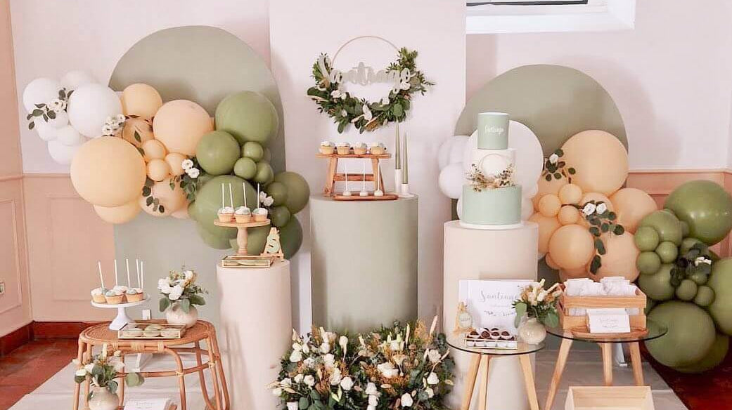 Decoração moderna de baby shower com balões, bolos e mesas elegantes em tons suaves de verde e bege.