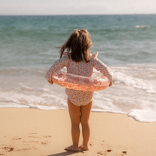 Child at the beach holding a pink flower-patterned floating ring, ready for summer fun.