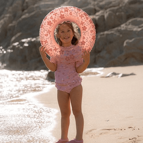 Little girl on the beach holding a pink floral swim ring from Little Dutch, enjoying summer fun by the water.