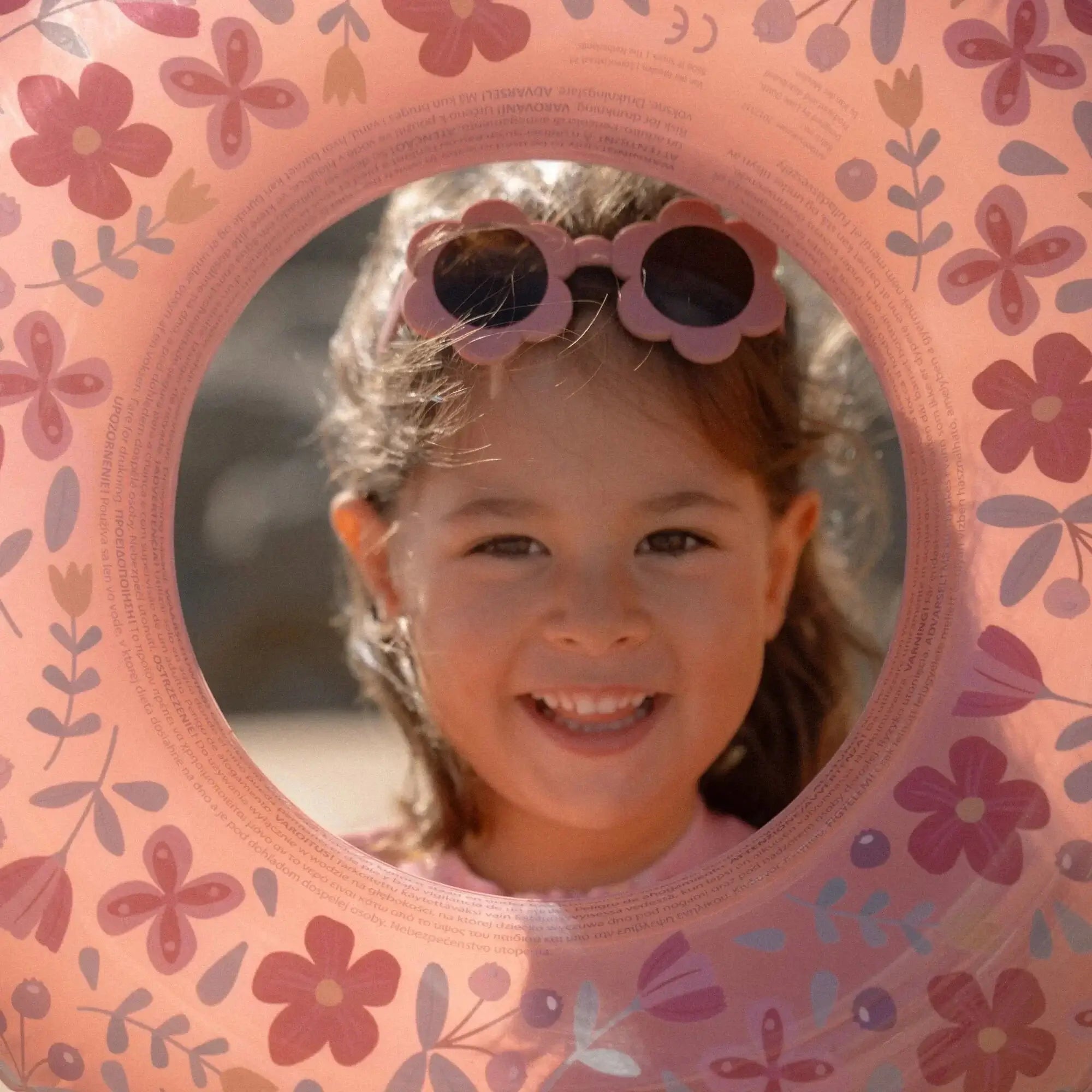 Girl wearing Flower Pink sunglasses by Little Dutch, smiling through a floral swim ring, perfect for sunny days.