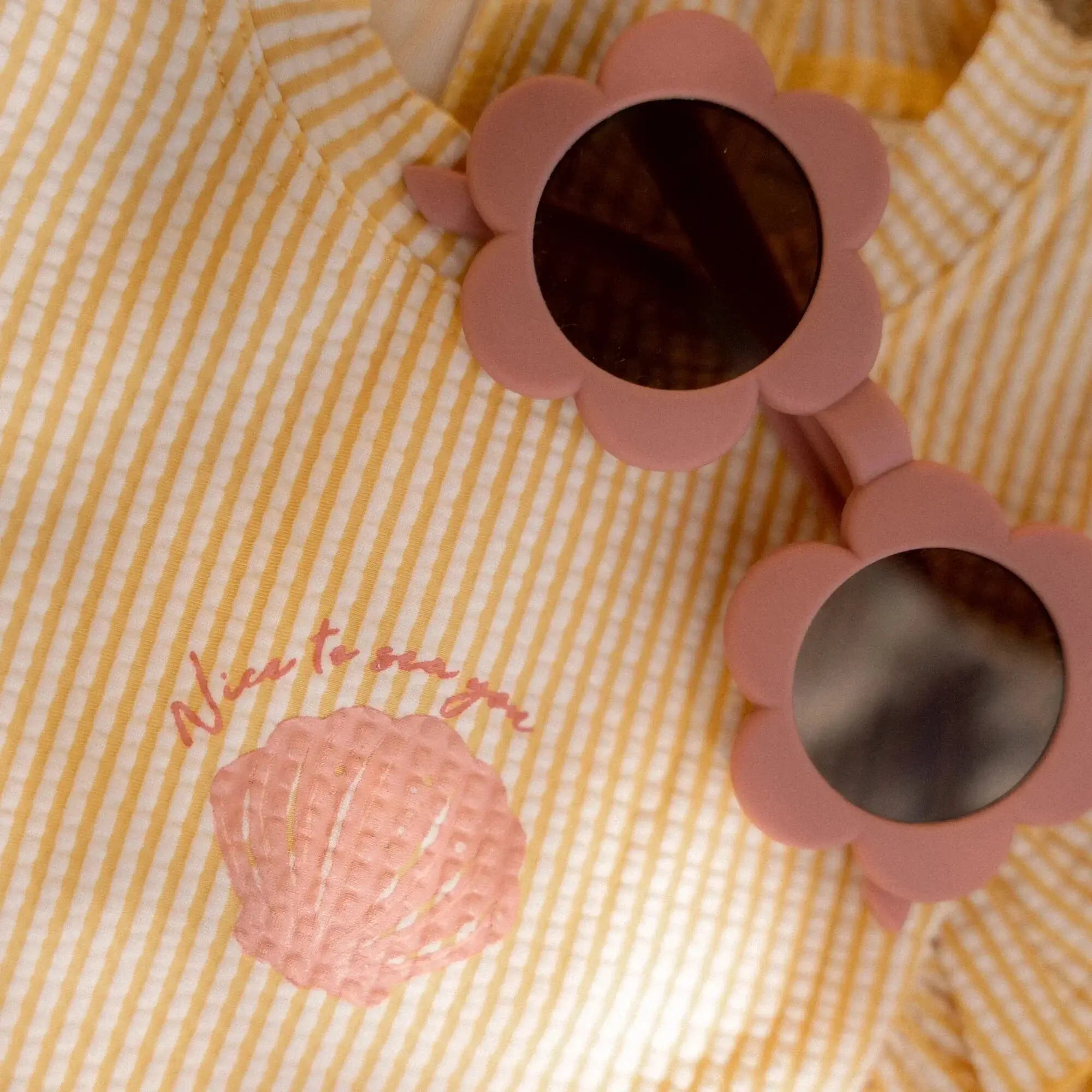 Óculos de sol para crianças em design de flor rosa, ao lado de uma blusa listrada amarela com concha.