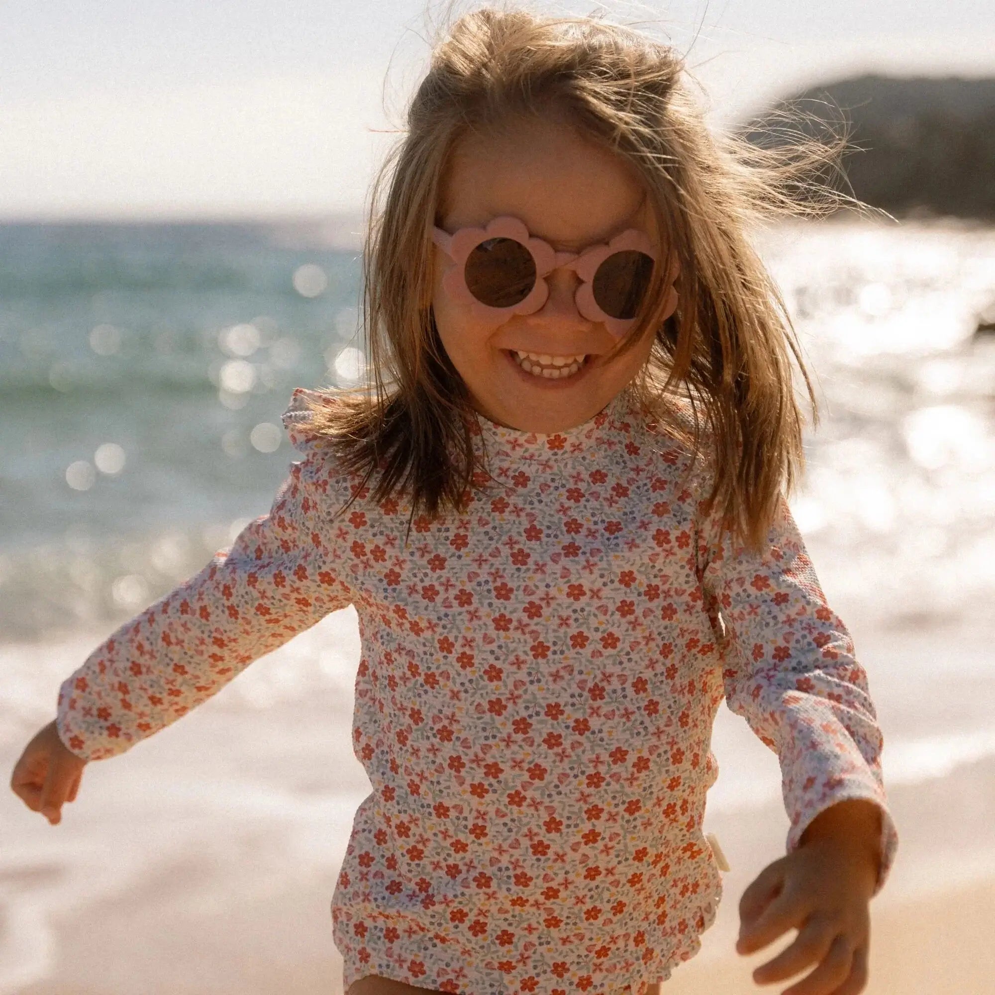 Criança alegre correndo na praia usando óculos de sol Flower Pink da Little Dutch, com proteção UV400.