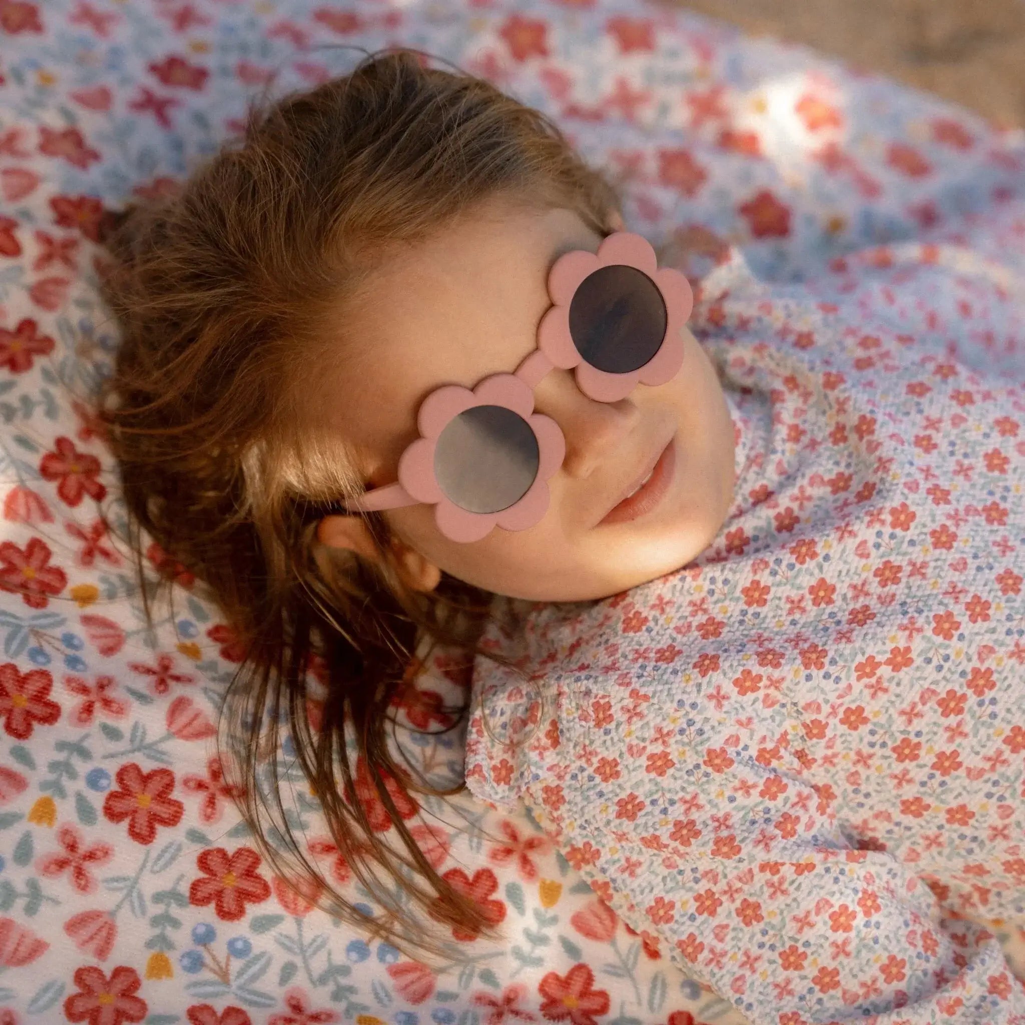 Criança usando óculos de sol Flower Pink Little Dutch, deitada em um cobertor floral, protegendo os olhos no sol.