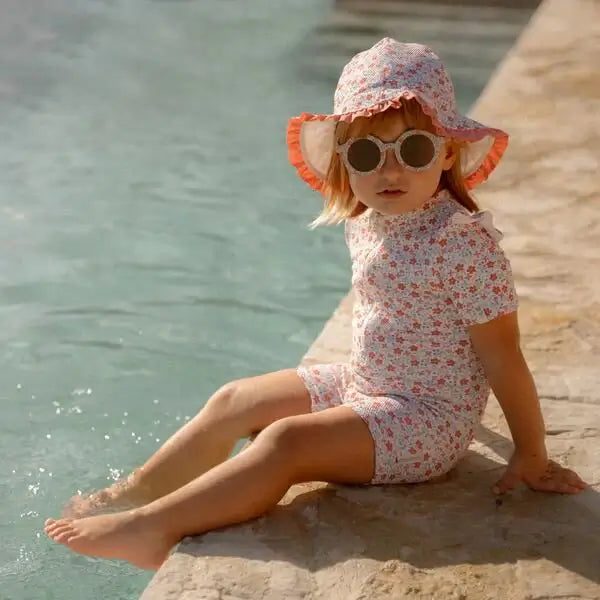 Criança usando óculos de sol redondos e chapéu florido, relaxando à beira da piscina em um dia ensolarado.