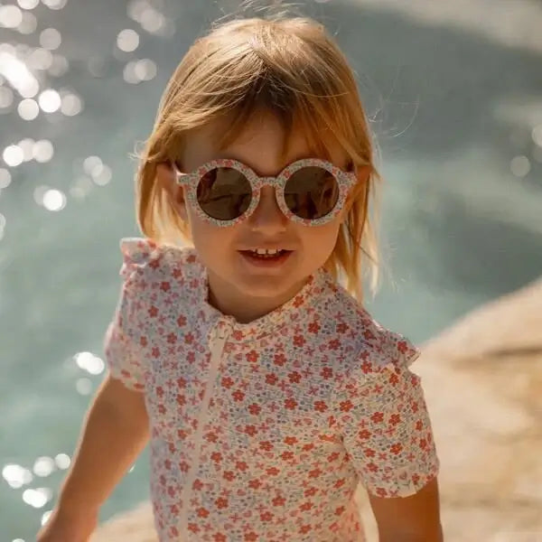 Criança usando óculos de sol redondos com estampa floral, aproveitando um dia ensolarado à beira da água.