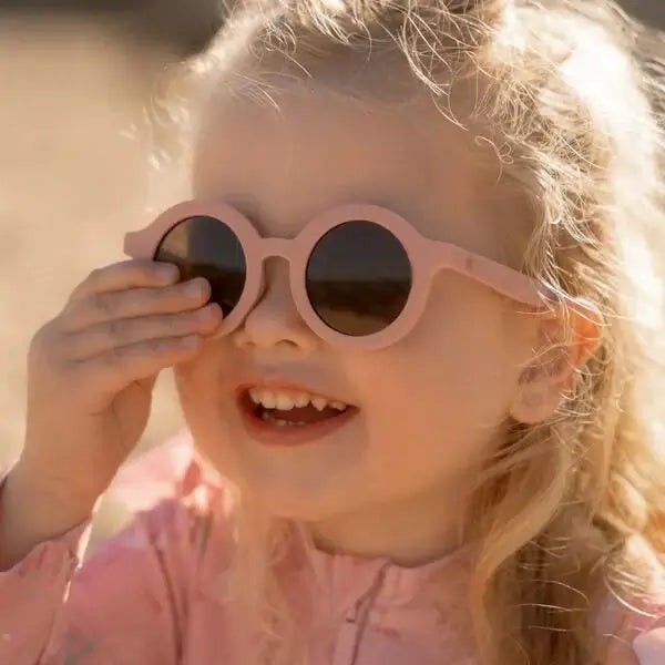 Criança sorridente usando óculos de sol de cor rosa claro, proteção UV para dias ensolarados.