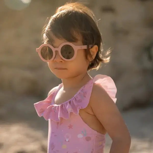 Criança usando óculos de sol redondos rosa com maiô rosa, ideal para proteção e estilo ao ar livre.
