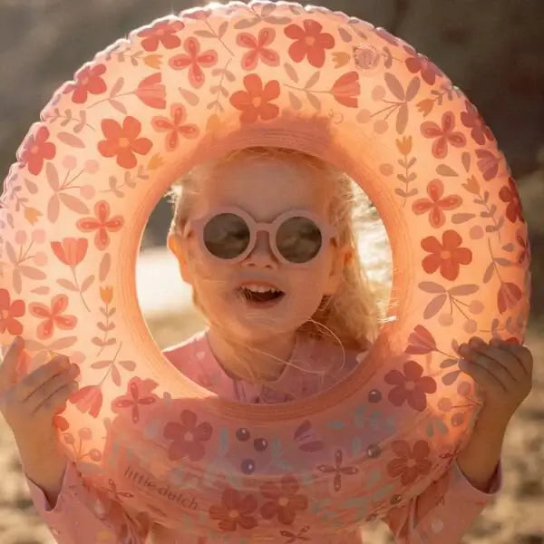 Criança sorridente usando óculos de sol redondos rosa, segurando boia com flores no dia ensolarado.