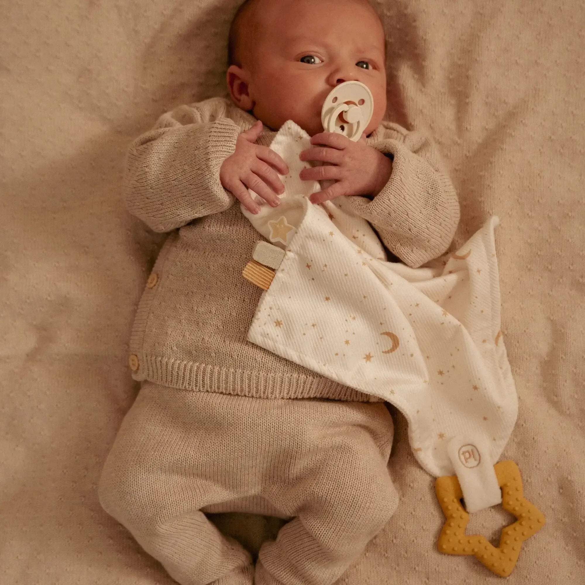 Bebé com doudou e anel de dentição, usando roupas aconchegantes e chupeta, em fundo suave.