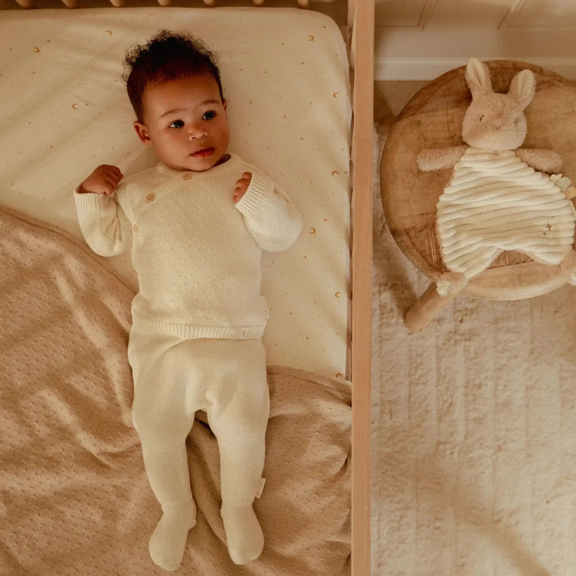 Newborn baby peacefully lying in crib with soft beige outfit, next to Doudou Coelho plush toy from Little Dutch.