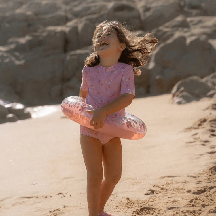 Young girl in pink swimsuit joyfully holding a Little Dutch pink flower inflatable pool ring on the beach.