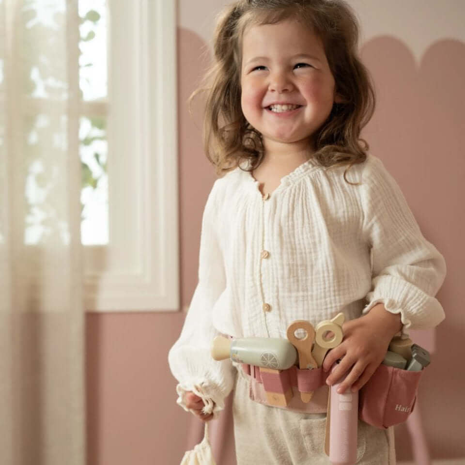 Garota sorridente usando o Conjunto de Cabeleireiro Little Dutch com acessórios em bolsa de cintura ajustável.