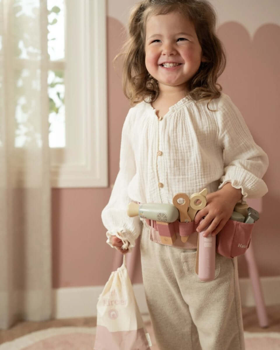 Garota sorridente usando o Conjunto de Cabeleireiro Little Dutch com acessórios em bolsa de cintura ajustável.