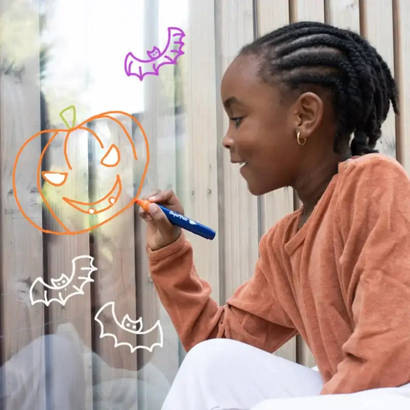 Criança desenhando em vidro com marcadores coloridos, incluindo um abóbora e morcegos, usando marcadores SuperPetit.