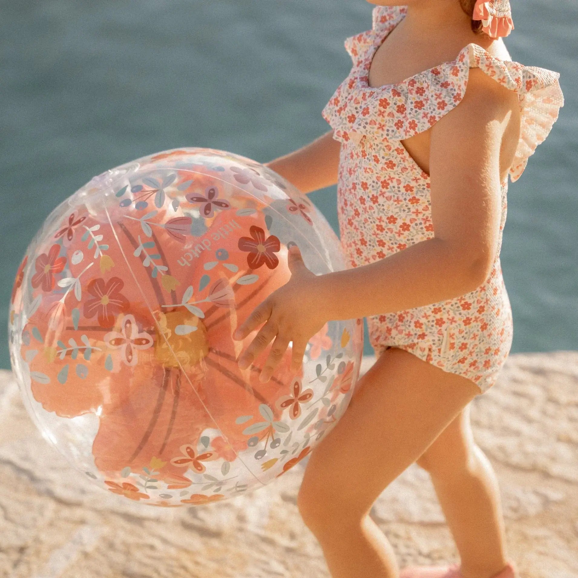 Criança segurando bola insuflável de praia 3D com flores rosas, à beira de uma água azul, vestindo maiô floral.