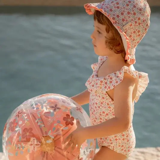 Child in a floral swimsuit holding a 3D inflatable beach ball with pink flowers by the water.
