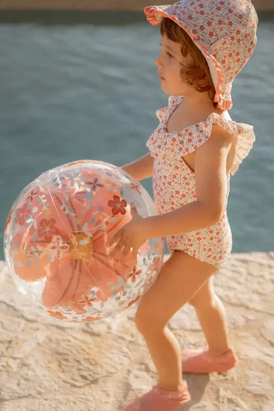 Child in a floral swimsuit holding a 3D inflatable beach ball with pink flowers by the water.
