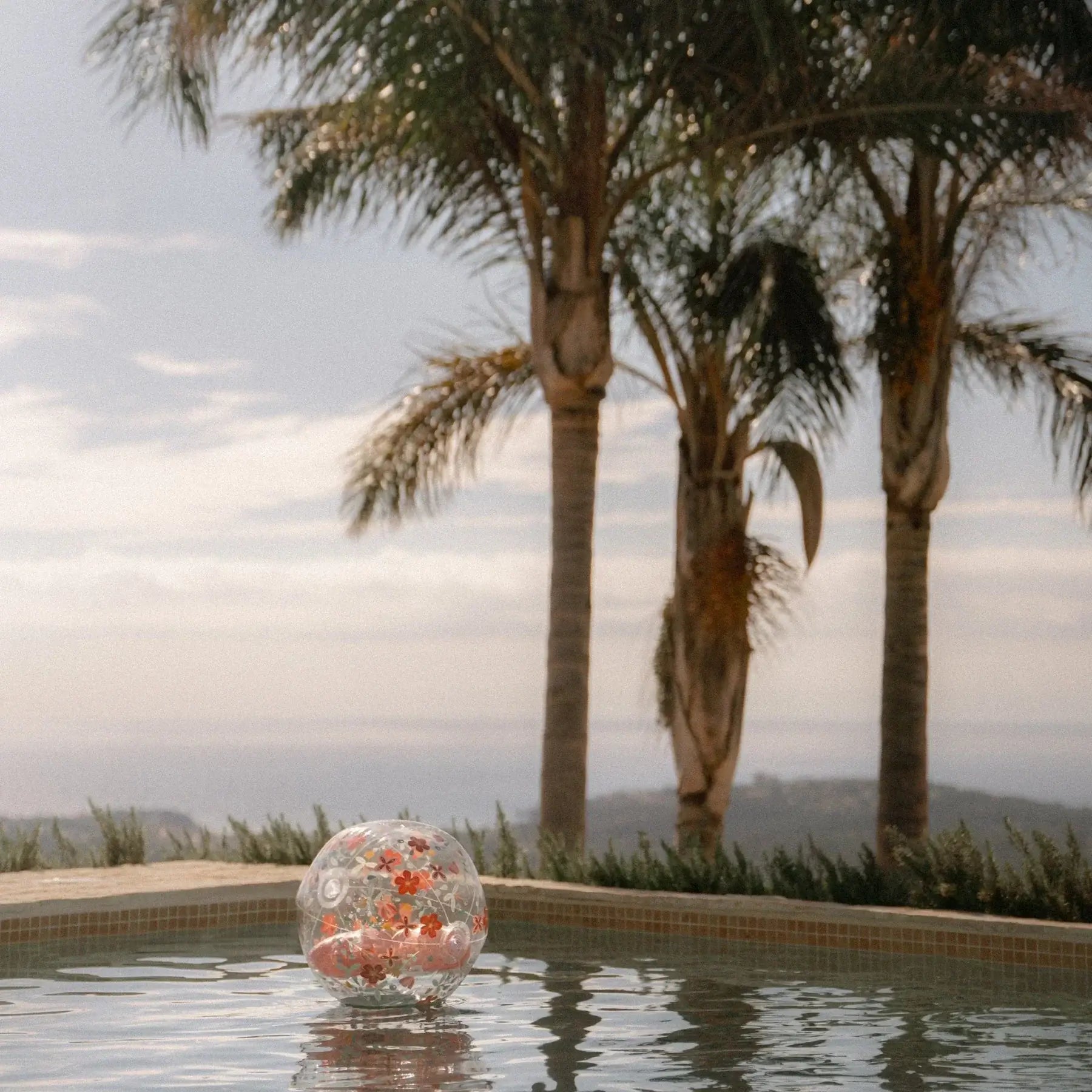 Bola insuflável de praia 3D com flores pink flutuando na piscina com palmeiras ao fundo.