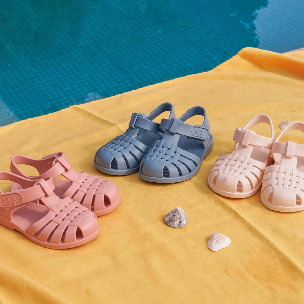 Igor children's sandals in pink, blue, and ivory displayed on a sunny yellow cloth near a pool.