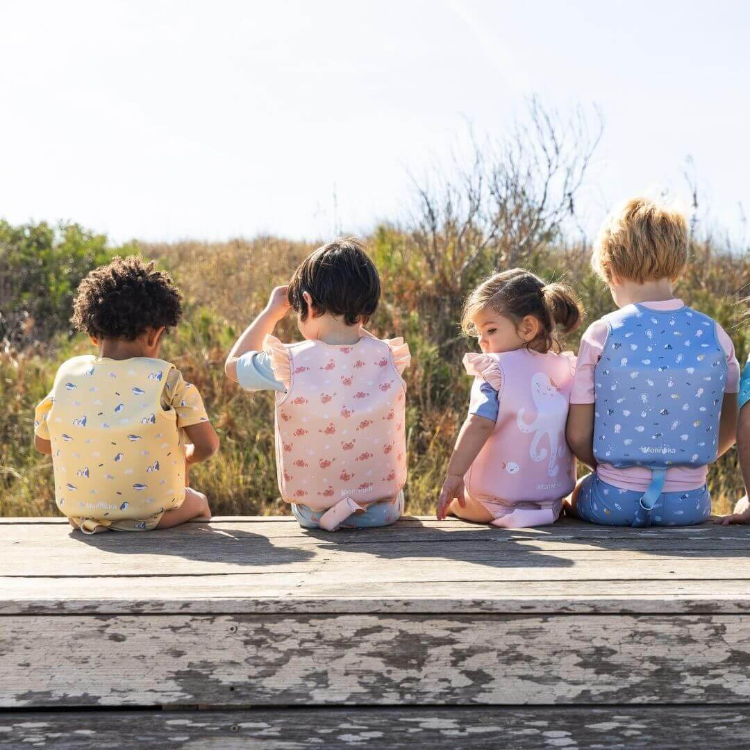 Crianças usando coletes flutuadores coloridos Monneka, sentadas em um deck ao ar livre em frente à natureza.