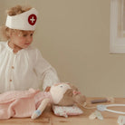 Child playing doctor with a doll, wearing a nurse's cap, surrounded by medical toys in a cozy setting.