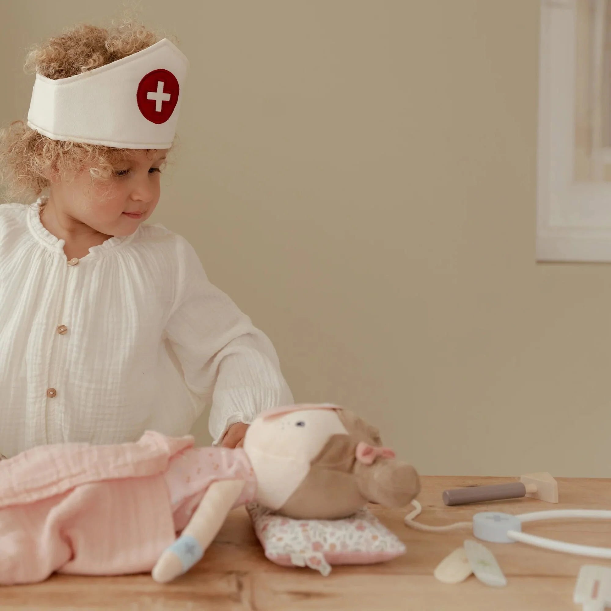 Child playing doctor with a doll, wearing a nurse's cap, surrounded by medical toys in a cozy setting.