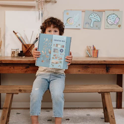 Child holding the Kit de Criatividade XL Forest Friends creative box, showcasing arts and crafts materials in a playful setting.