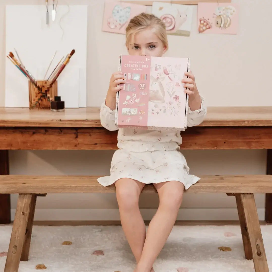 Girl holding Little Dutch XL Fairy Garden creativity kit while seated at a wooden table, surrounded by craft supplies.