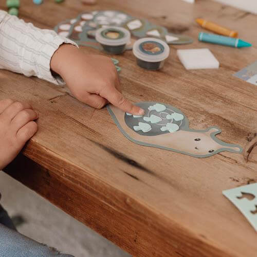 Child's hand touching a snail craft project surrounded by colorful crafting materials on a wooden table.