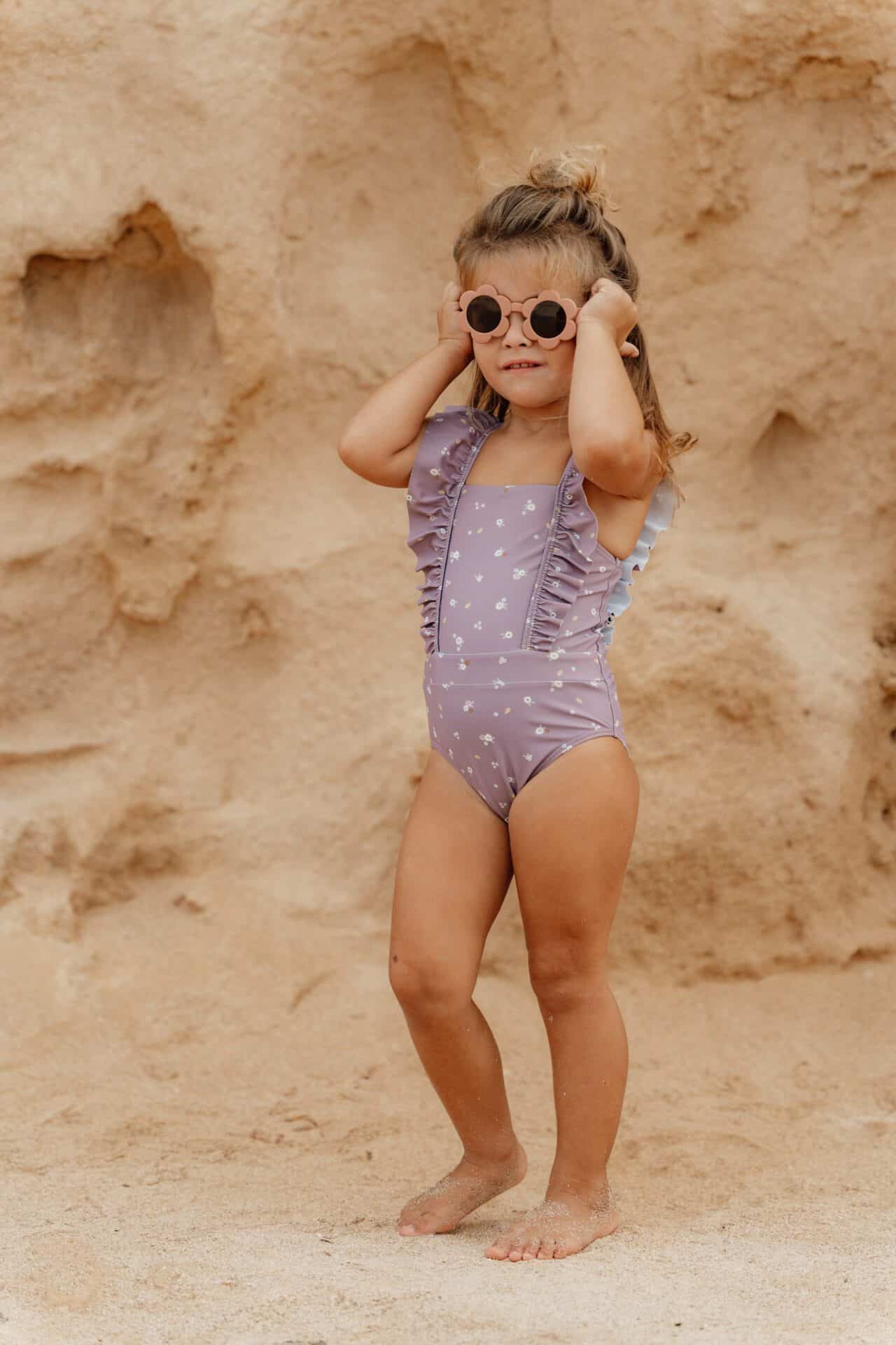 Child wearing Little Dutch mauve swimsuit with straps, standing on sandy beach with rocky background