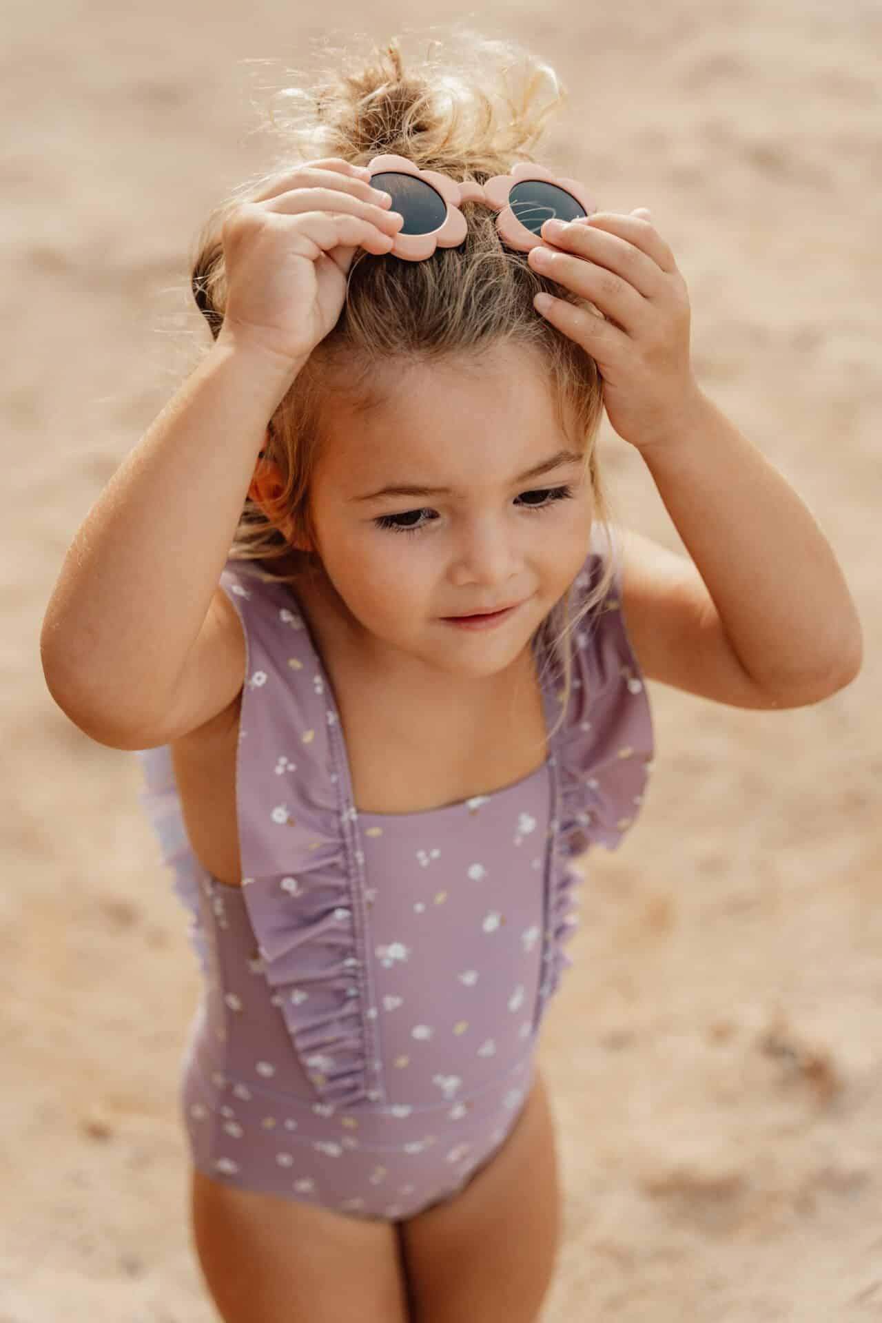 Little girl wearing mauve Little Dutch swimsuit with shoulder straps and adjusting sunglasses at the beach.