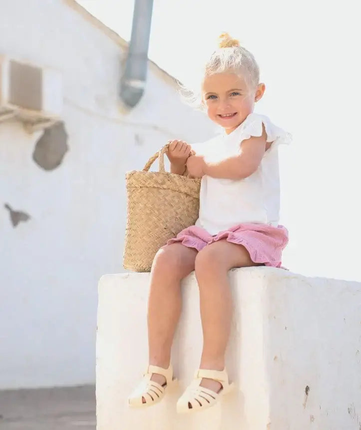 Criança sorridente usando sandálias Igor tamanho 23, segurando uma cesta de palha, em um ambiente de verão.
