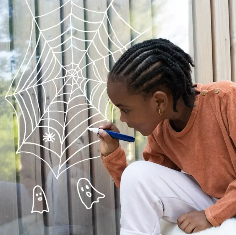 Criança desenhando uma teia de aranha com marcadores de vidro em uma janela, estimulando a criatividade infantil.