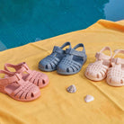 Igor children's sandals in pink, blue, and ivory displayed on a sunny yellow cloth near a pool.