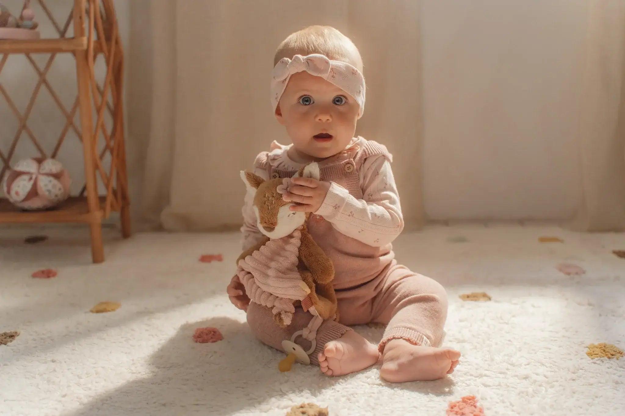 Baby sitting on a soft rug, holding a Little Dutch Fairy Garden Doudou corça, wearing a pink headband and outfit.