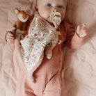 Baby holding a soft doudou with a deer design, featuring a pacifier and floral patterns on a cozy blanket.