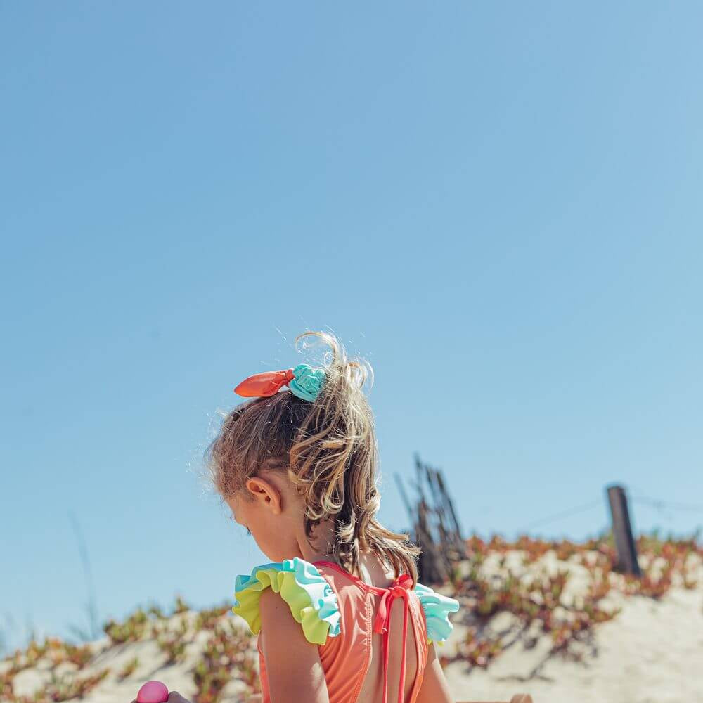 Menina com fato de banho Candy Colors, de costas em uma praia ensolarada, com design de laço nas costas e babados coloridos.