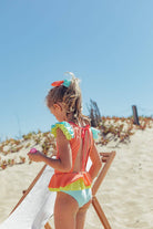 Menina com fato de banho Candy Colors, de costas em uma praia ensolarada, com design de laço nas costas e babados coloridos.