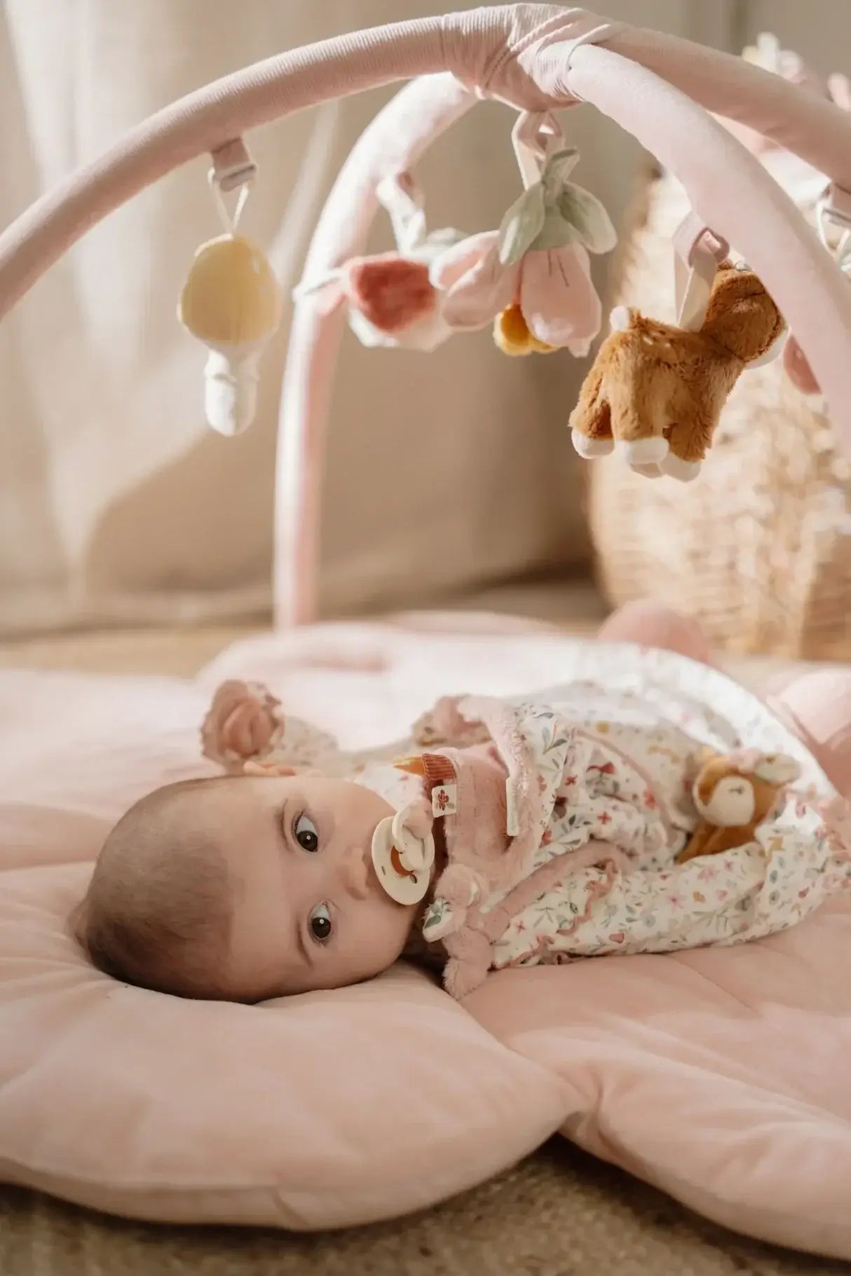 Baby playing under the pink activity gym with toys on the soft floral-patterned mat.