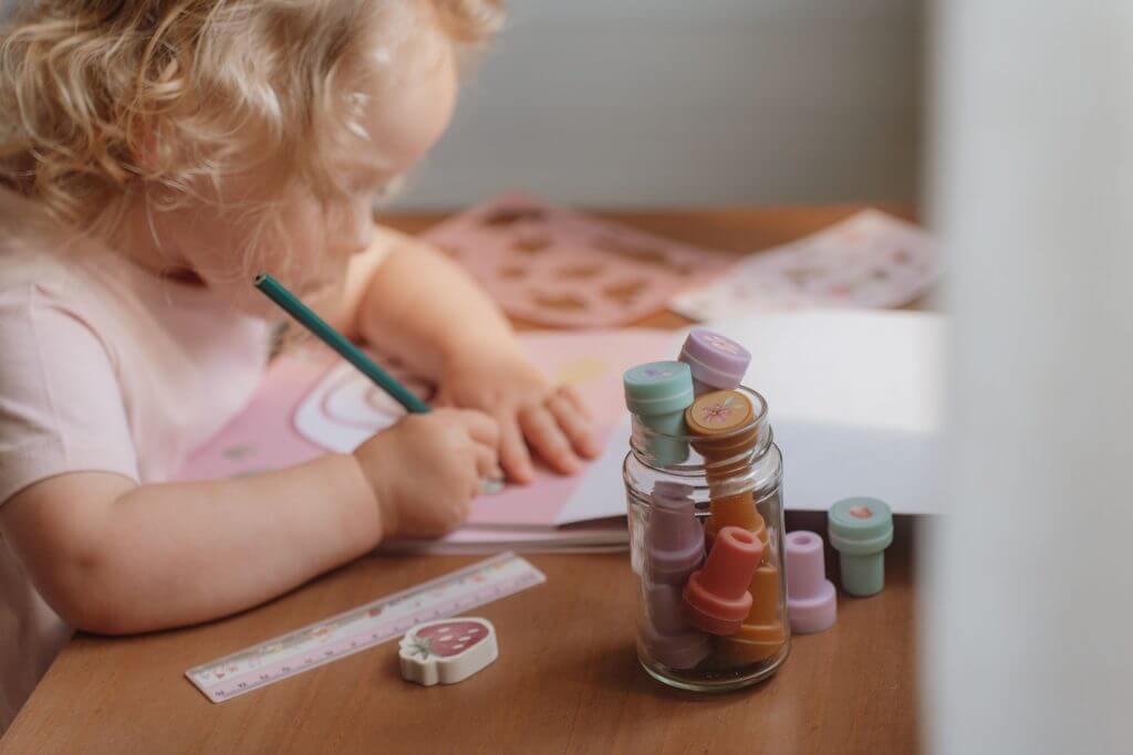 Criança colorindo com lápis enquanto um pote de carimbos coloridos está ao lado em uma mesa de madeira.