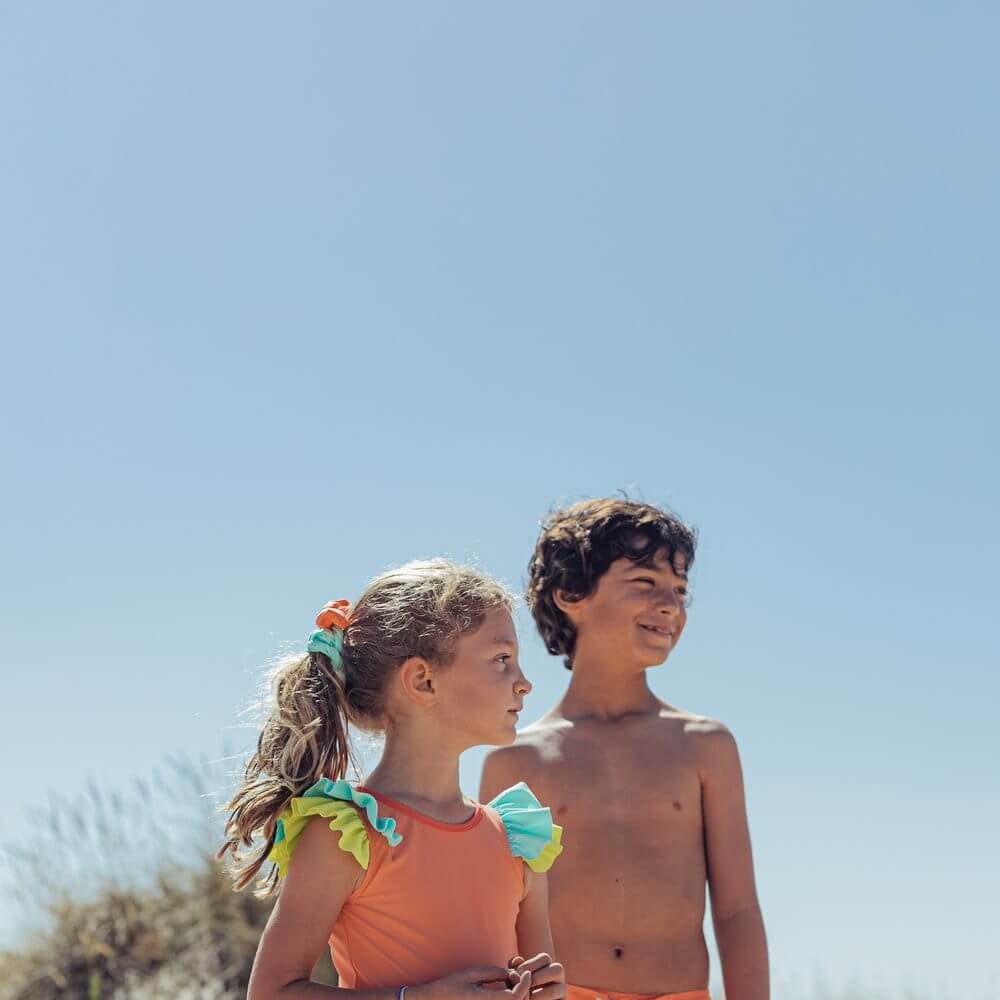 Menina e menino na praia, vestindo fato de banho de candy colors da Paper Boat, aproveitando um dia ensolarado e divertido de Verão.