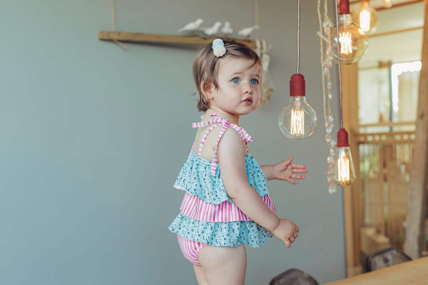 Criança usando fato de banho com folhos, padrão de folhas verdes e riscas rosa, alças ajustáveis com lacinhos nos ombros