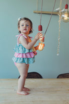 Child wearing Green Leafs swimsuit with frills and pink stripes, standing on table holding light bulb.
