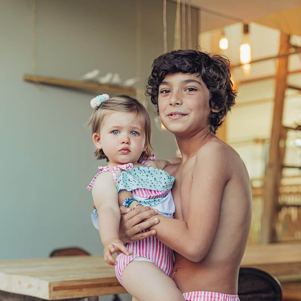 Child wearing Green Leafs swimsuit with green leaf pattern and pink stripes, being held by an older sibling in matching swim shorts.