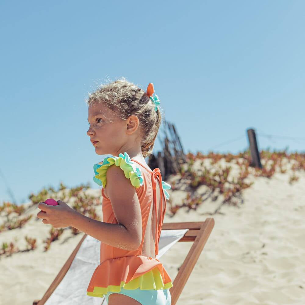 Menina usando fato de banho de Candy Colors da Paper Boat, com design colorido e laço nas costas, no verão na praia.