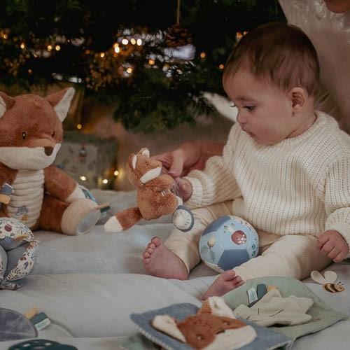 Bebé brincando com brinquedos em um tapete acolchoado perto de uma árvore de Natal, promovendo exploração e diversão.